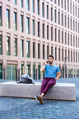 Portrait of cheerful modern businessman speaking by phone and smiling while sitting outdoors in a bench