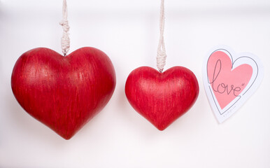 Two red hanging love hearts, one big on the left and a smaller one on the right, plus a paper heart with love written on it, on a white background