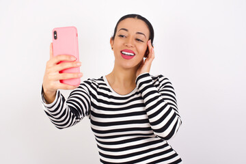 Young beautiful woman wearing stripped t-shirt against white background smiling and taking a selfie...
