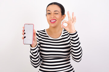 Young beautiful woman wearing stripped t-shirt against white background holding in hands cell...