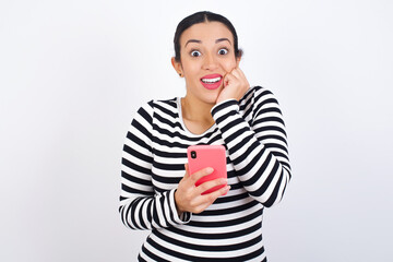 Joyous Young beautiful woman wearing stripped t-shirt ag poses with mobile phone device, types text message on modern smartphone, watches funny video during free time, enjoys good internet connection,