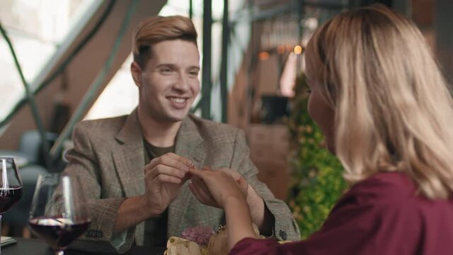Medium over-the-shoulder footage of smiling man wearing ring on his girlfriend hand making proposal sitting at table in restaurant