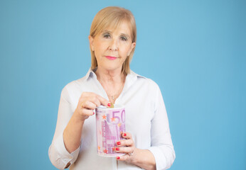 Senior woman puts money into the piggy bank over blue background