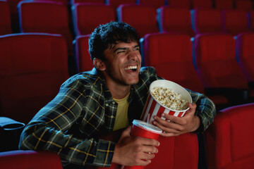 Portrait of joyful young guy laughing, holding a drink and popcorn basket while watching movie...