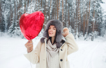 Portrait of a young girl with a bright heart-shaped balloon in a snow-covered forest. Valentine's Day