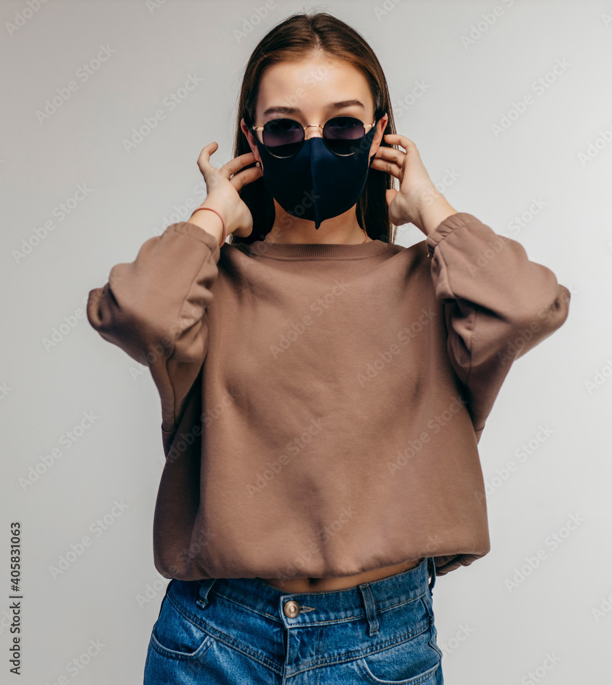 Wall mural Young woman in glasses, medical mask and hoodie posing in studio on white background - Wall murals