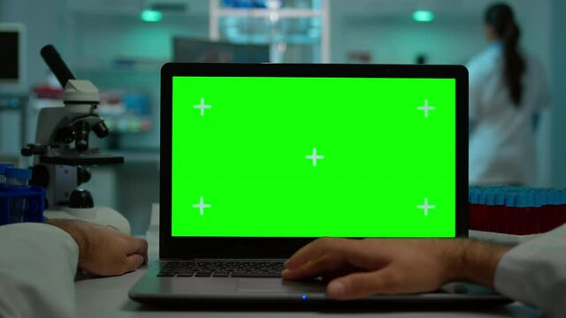 Pov shot of man scientist sitting at desk working on laptop with mock-up green screen, isolated display. In background lab researcher analysing vaccine developent examining samples
