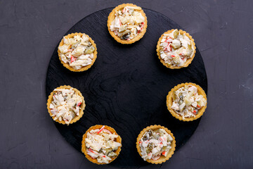 Tartlets with salad with pineapple and shrimps on a black wooden round board on a gray concrete background copy space