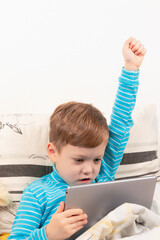 child boy in a blue sweater at home in bed plays on the tablet on the background of a white wall