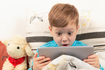 child boy in a blue sweater at home in bed plays on the tablet on the background of a white wall