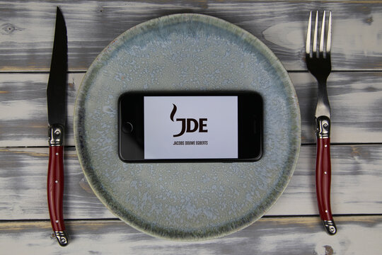 Viersen, Germany - May 9. 2020: Closeup of mobile phone on plate and wood table, cutlery with logo lettering of jde jacobs douwe egberts coffee food company (focus on center)