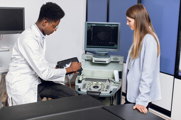 Young male african doctor in modern clinic, holding tablet pc and showing to his female caucasian patient ultrasound scan of kidneys and internal organs and explaining ways of treatment.