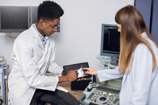 Professional African man ultrasound specialist, holding ipad pc with ultrasound scan image and explaining ways of treatment kidney stones for his young female Caucasian patient