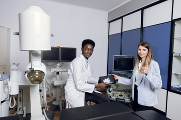 Portrait of smiling African man doctor, holding digital tablet with ultrasound scan, posing to camera, while consulting his female patient in modern clinic. Woman patient shows thumb up