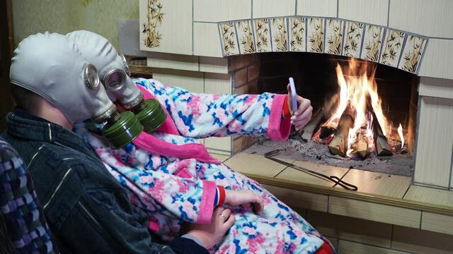 Epidemic, Covid-19. A woman and a man during the pandemic sit near the fireplace in gas masks and take selfies on the phone.