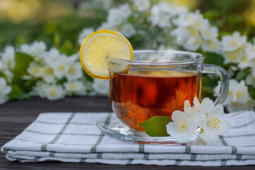 Glass cup with tea, lemon, napkin, jasmine branches on a wooden table. Outdoor, picnic, brunch. Selective focus with short DOF. Close up view.