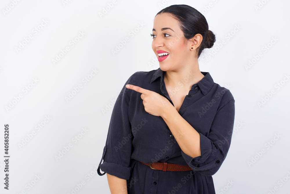 Wall mural Happy cheerful smiling young beautiful Arab woman wearing gray dress against white studio wall looking and pointing aside with hand. Copy space and advertisement concept.