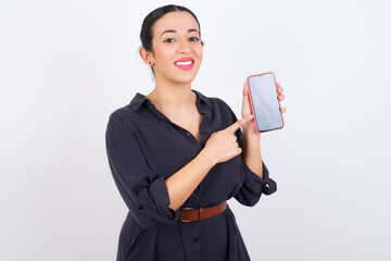 Smiling young beautiful Arab woman wearing gray dress against white studio background showing and pointing at empty phone screen. Advertisement and communication concept.