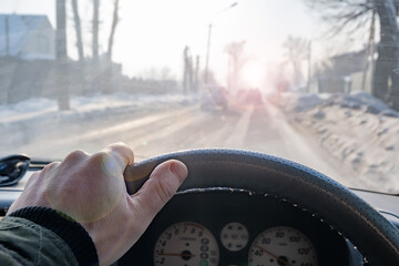 a dangerous road, a man's hand on the steering wheel of a car that is driving on a snow covered slippery icy winter road and the sun's glare impairs the visibility of the path, blinding the eyes