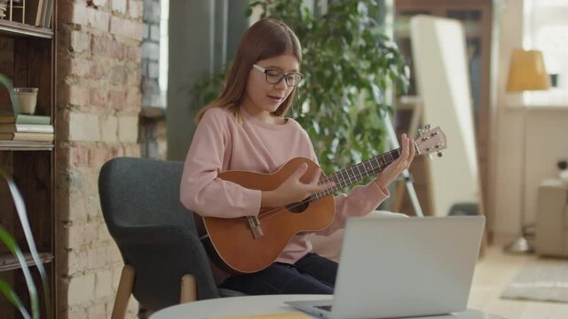 Medium shot of 12-year-old girl sitting on chair at home having online music lesson on laptop practicing playing ukulele