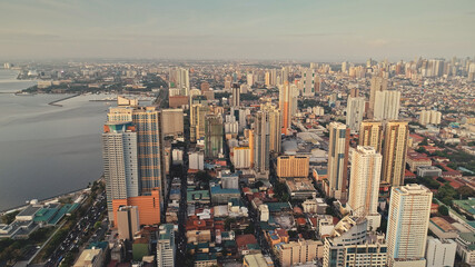 Fototapeta premium Modern cityscape at ocean bay aerial. High scyscrapers and block buildings at sea coast. Traffic highway with cars, trucks. Green park at roadside. Cinematic capital town of Manila, Philippines, Asia