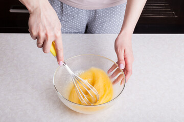 A woman in the kitchen holds a whisk in her hands with a yellow handle and whips them with eggs in a glass transparent bowl. Mother’s daily routine - making fried eggs breakfast for the family.