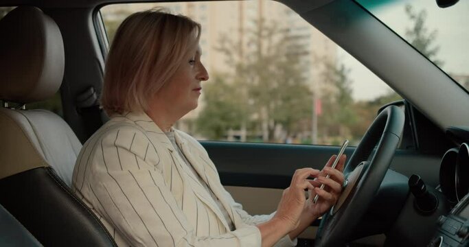 Aged female in business suit scrolling smartphone, sitting in car, technology