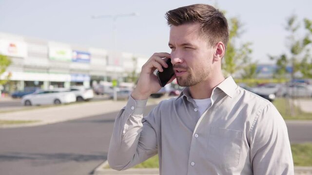 A young Caucasian man talks on a smartphone in an urban area - a shopping center and a parking lot in the blurry background