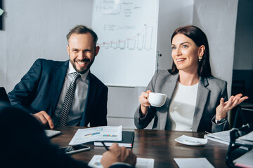 Cheerful businesspeople with coffee talking with advisor on blurred foreground near papers on table