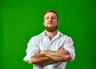 A Caucasian with a beard folded his hands in a white shirt. A muscular man on a green background with strong arms. High quality photo