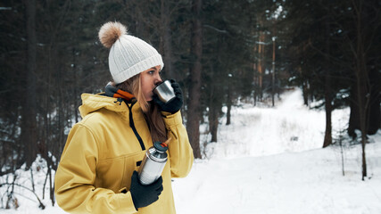 Fototapeta na wymiar A girl in a yellow jacket drinks tea from a thermos in the winter in the forest