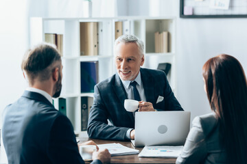 Smiling investor with cup of coffee looking at businesspeople on blurred foreground near papers and laptop