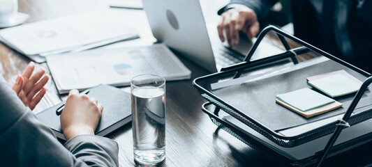 Cropped view of businesswoman holding pen near notebook and glass of water during consultation with investor on blurred background, banner