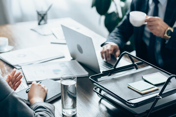 Naklejka premium Cropped view of businesswoman holding pen near notebook, glass of water and investor with cup of coffee in office