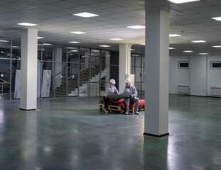 A man and woman in work clothes and hard hats sit on pallets and look at blueprints in a large empty room.
