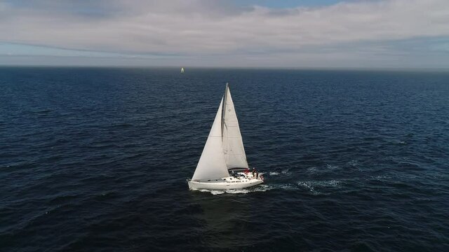 Aerial view. A white sailboat is moving fast on the waves of the sea. Flying around a boat in full sailing gear. Close hauled sailing in ocean. Yacht in windy and sunny day. Drone shot.