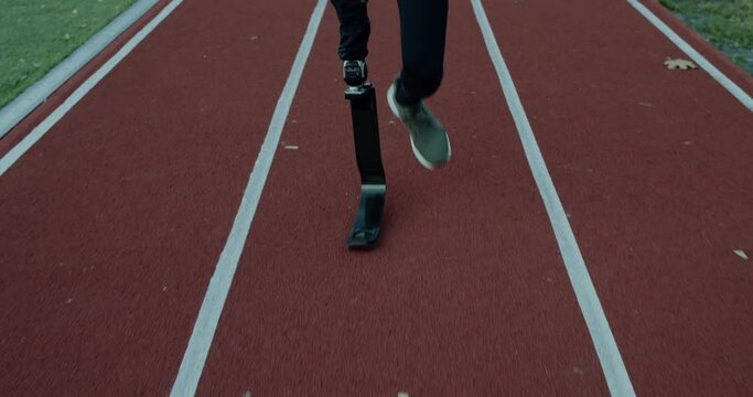 Crop view of unknown disabled male person with prosthetic blades running at sports field. Amputee male sportsman jogging. Concept of motivational sports footage.