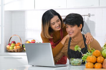 Young Asian couple eating salad while using laptop at table in kitchen . Eating healthy Vegetarian food . 