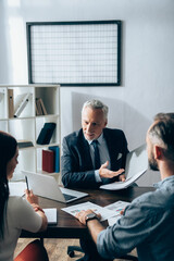 Obraz premium Smiling investor pointing with hand at paper folder near businesspeople on blurred foreground and laptop on table