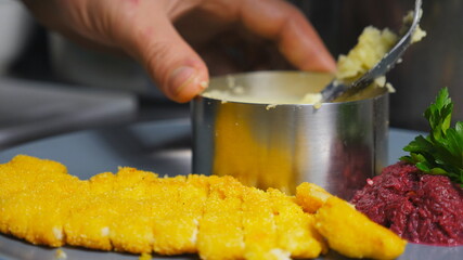 Close up male hand of cook putting mashed potatoes with the spoon into the iron mold at kitchen. Chef is serving puree with breaded chicken fillet on plate. Concept of preparing food. Slow motion