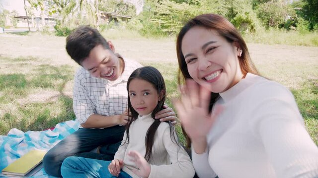 Happy family picnic. Asian parents (Father, Mother) and little girl take a selfie by smartphone with have fun and enjoy ourselves together during picnicking on a picnic cloth in the green garden