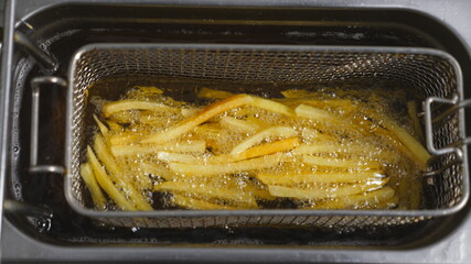 Top view of male cook shaking a lattice with french fries being prepared in hot boiling oil. Chef lowering a grid with potatoes into deep fryer. Process of making fast food at cuisine. Slow motion