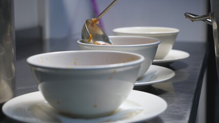 Male cook pouring red beetroot soup from pan in white bowl using a ladle at kitchen. Chef preparing to serve traditional ukrainian and russian borscht at restaurant. Concept of cooking food. Close up