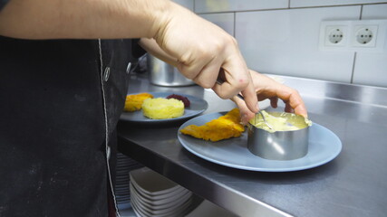 Male hand of cook putting puree with the spoon into the iron mold at kitchen. Chef is serving mashed potatoes with breaded chicken fillet on plate. Concept of preparing food. Dolly shot Slow motion