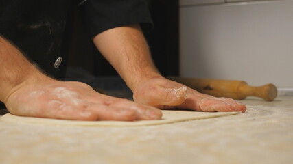 Obraz na płótnie Canvas Male arms of cook forming pastry on a wooden surface at restaurant. Hands of chef shaping floured dough for pizza on a kitchen table at cuisine. Concept of preparing food. Close up Slow motion