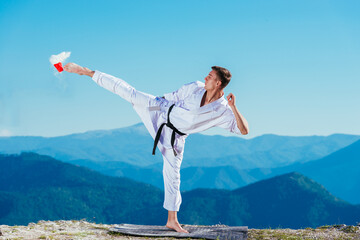 Fit karate athlete kicking a cup filled whit fluor causing a big splash while wearing a white kimono on top of a mountain on a sunny day. © qunica.com