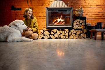 Woman sitting by the fireplace with a white dog at cozy and loft style home interior - obrazy, fototapety, plakaty