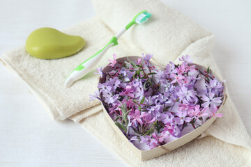 Obraz na płótnie Canvas Packaging in the form of a heart. filled with delicate purple flowers on the background of a toothbrush and soap on a white bath towel - a concept of pleasant self-care