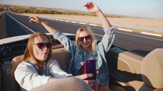 Two beautiful women ride in back seat of convertible and shoot video on their phone, smiling and laughing. Young women in sunglasses take selfies while ride car without a roof