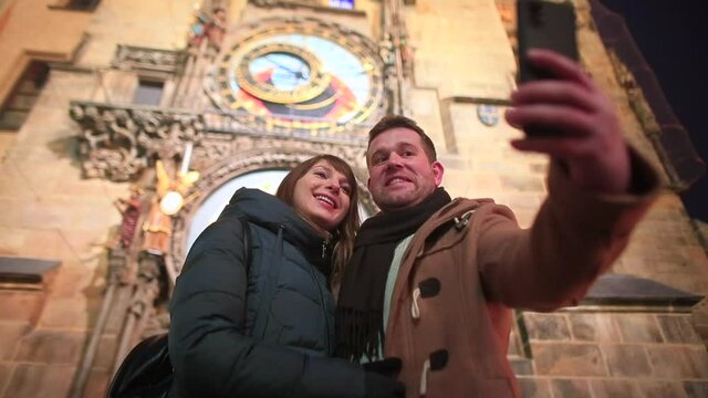 A man uses phone in hands, makes selfie video. They are posing in old town of Prague, against background clock. Exchange of emotions and photos on a social network using the mobile Internet.